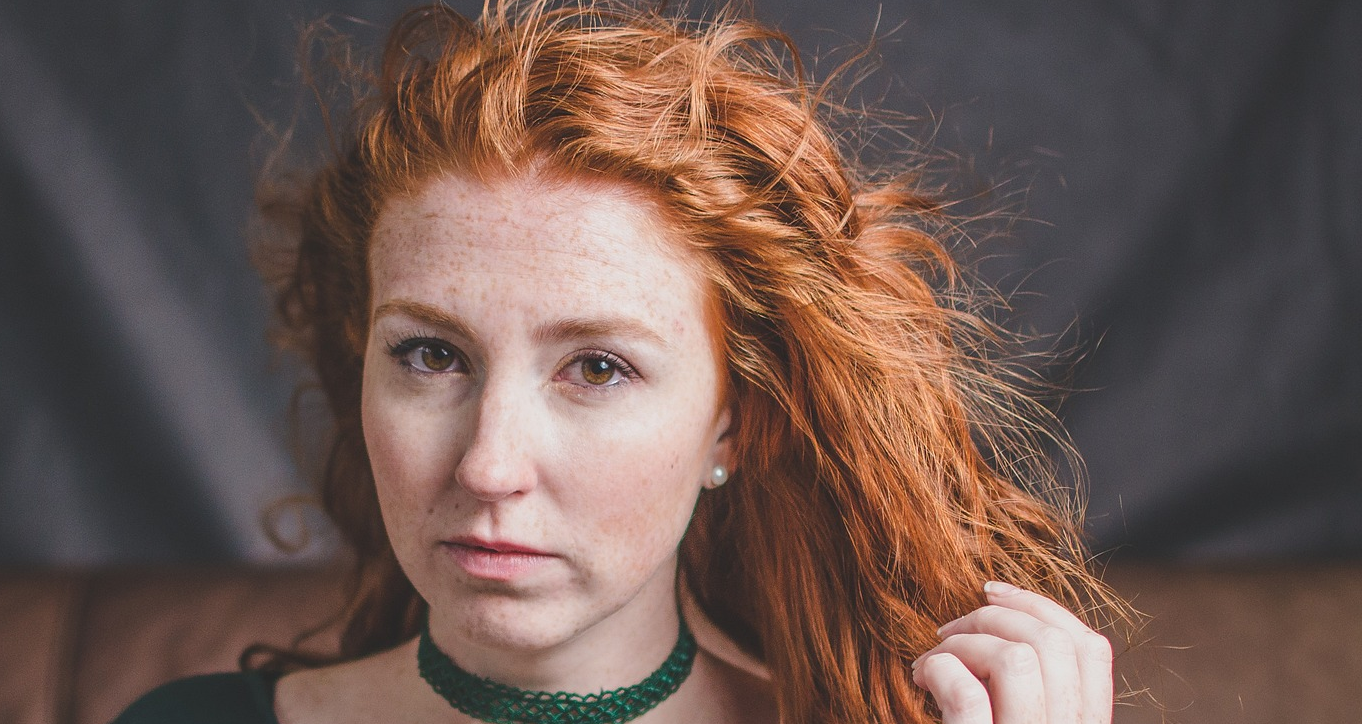 Moira from Meadowsweet. Woman with pale skin, wavey red hair, wearing a green laced top and matching choker.